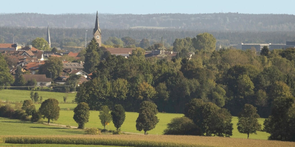 Blick auf die Stadt Holzkirchen mit Kirchtürmen und roten Dächern, umgeben von grünen Feldern und Wäldern – moderne Infrastruktur trifft auf naturnahe Lebensqualität.