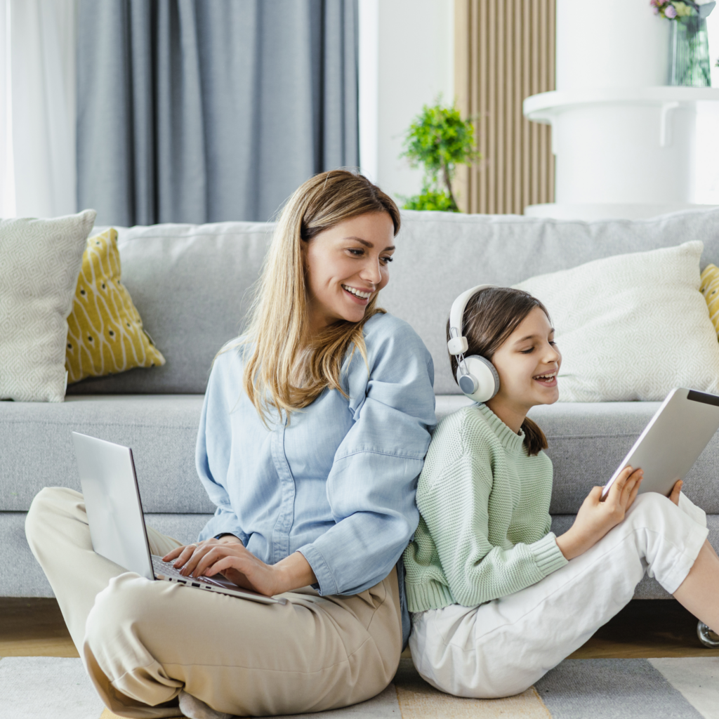 Moderne Internetnutzung im Homeoffice. Eine Mutter, am Notebook arbeitetend, sitzt Rücken an Rücken mit ihrer Tochter, die über Bluetooth Kopfhörer Musik hört und ein digitales Tablet in der Hand hält.